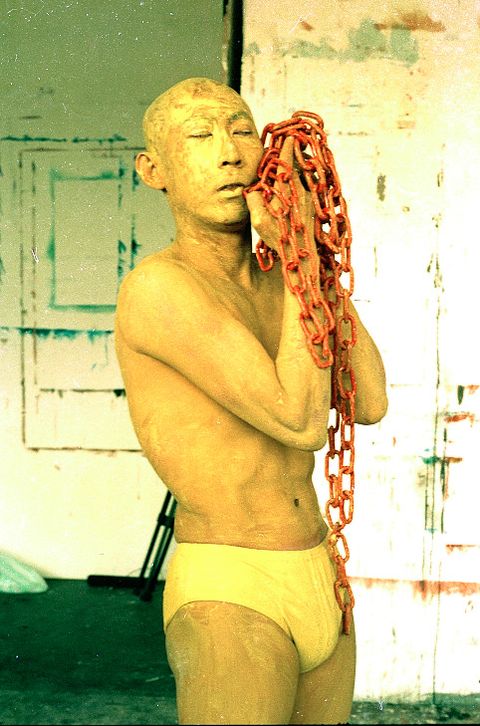 Performance still. The skin of a bald man wearing only yellow briefs is painted yellow. He holds up a handful of thick, red chains by his face, his eyes closed.