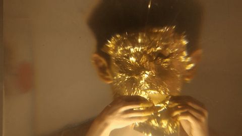 Video gif. A man applies gold leaf to his face in a repeating loop.