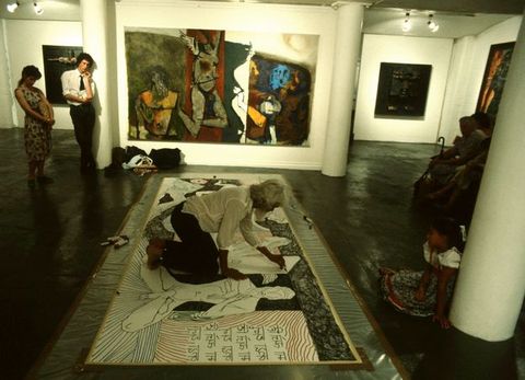 Installation. A person draws on a work of art spread on the floor at the centre of a gallery hung with paintings. The artist has brown skin, white hair, and is barefoot.