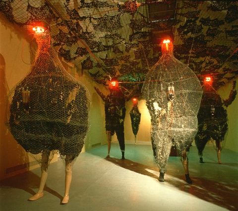 Installation. Five sculptural forms stand in a long gallery space. Each has the legs of a mannequin and bodies that are either humanoid or shaped more like a chicken. Each body has black-coloured material encased in chicken wire, and red lights glow like eyes from the very tops of tiny heads.