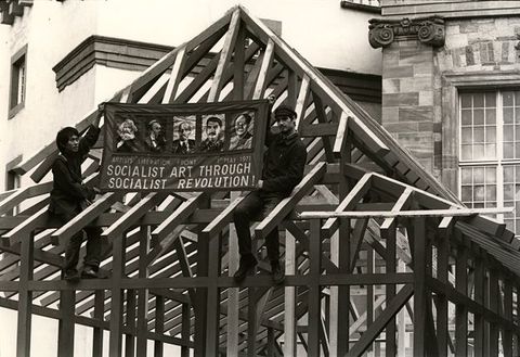 Black and white photograph. Two men hold up a sign reading “Socialist Art through Socialist Revolution!” The men are braced within the rafters of an exposed roof.