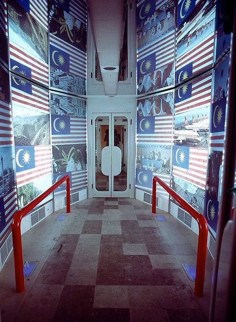 Installation. A walkway flanked by red railings lead to double doors. The walls are covered with the Malaysian flag alternating with photographs.