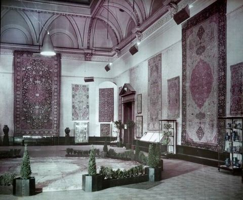 Colour transparency. Rugs are hung closely together on stone walls under elaborate moulding. The ceiling has lunettes set into curving arches lined with gilded carving. Two rugs reach nearly from the floor to the moulding around the high ceiling.