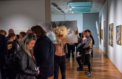 Performance still. A medium-skinned holds a sheet of magnifying plastic, arms stretched wide, as he walks towards us a museum. His face is covered in gold leaf, and his mouth is filled with red chili peppers. The magnifying material distorts the gold, making it look like rays of light. People crowd the space.