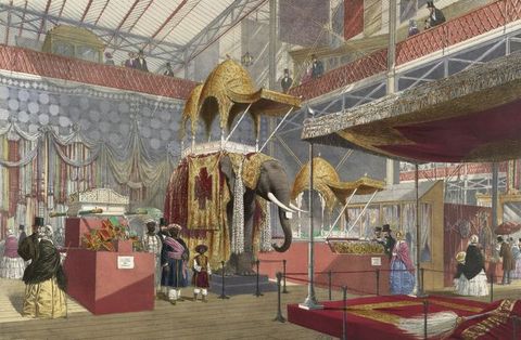 Illustration. A cavernous space has a taxidermy elephant at the centre. The animal is draped in red and gold cloth below a covered structure called a howdah. Two adults and one child with dark skin walk near the elephant. Pale-skinned men and women move around the exhibition or look down from a balcony level above.