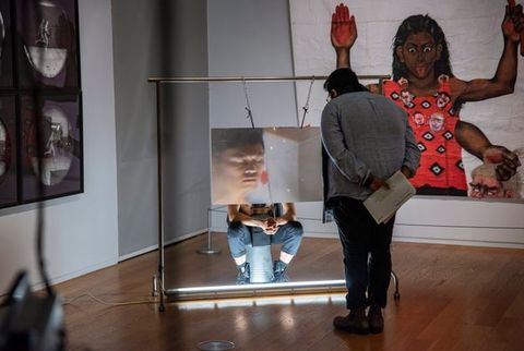 Installation. A person stands looking at a performance art piece. The artist sits on an upturned metal bucket with a magnifying sheet of plastic, the size of a large TV screen, which hangs from a metal frame down in front of his face. The artist’s eyes are closed and fingers interlaced, elbows resting on knees.